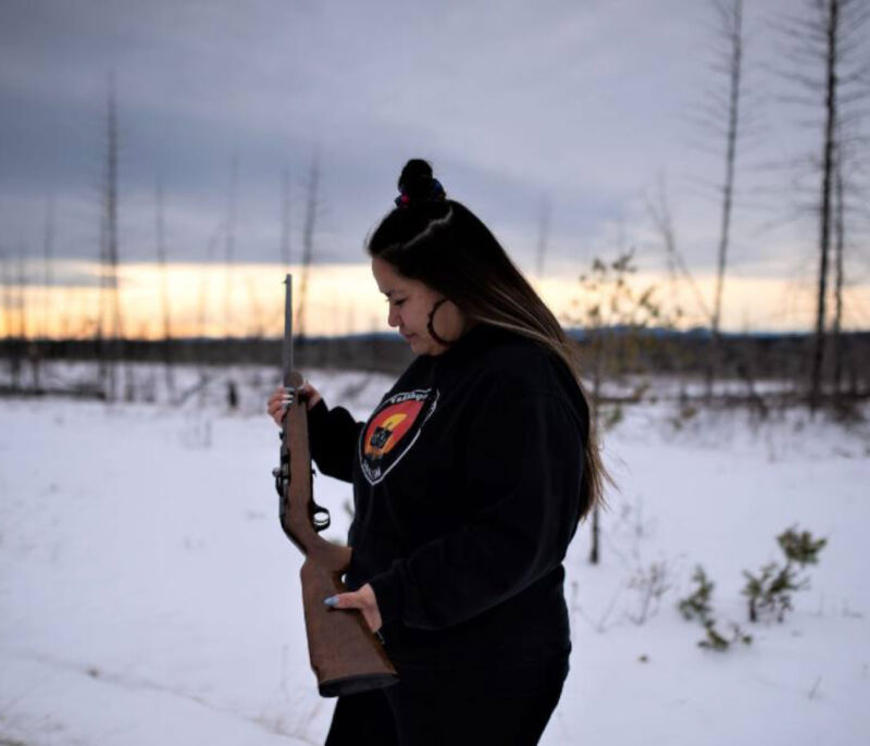 A woman with dark hair and hoop earrings stands in a snowing clearing holding a brown rifle.