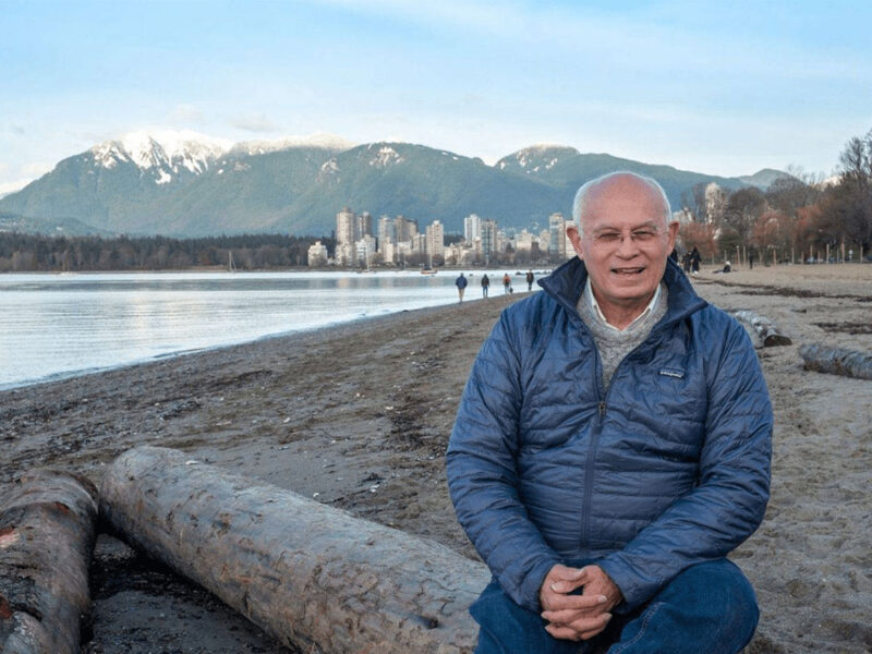 A photo of Wayne Crookes wearing glasses and a blue jacket, sitting on a log on a beach with a city & mountains in the distance.