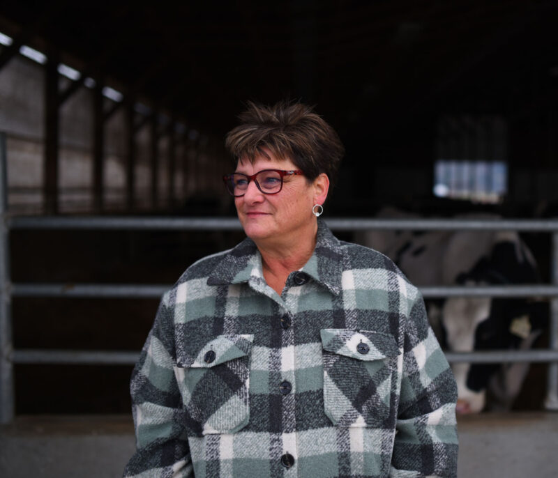A photo of Alison Arends wearing a plaid coat and glasses in front of a barn of cows and looking off into the distance.