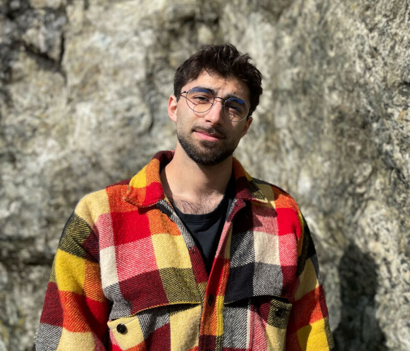 A photo of Tosh Sherkat in front of a large rock wearing glasses and a bright plaid shirt.